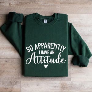 K&ouml;nnte beinhalten: Ein dunkelgr&uuml;ner Pullover mit dem Text "So Apparently I Have An Attitude" in wei&szlig;en Buchstaben. Ein kleines wei&szlig;es Herz befindet sich unter dem Text.