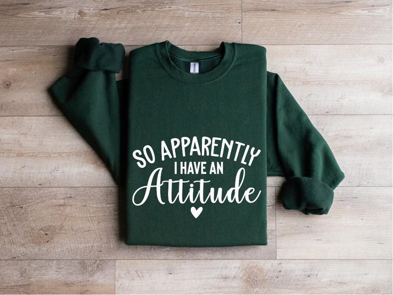 K&ouml;nnte beinhalten: Ein dunkelgr&uuml;ner Pullover mit dem Text "So Apparently I Have An Attitude" in wei&szlig;en Buchstaben. Ein kleines wei&szlig;es Herz befindet sich unter dem Text.