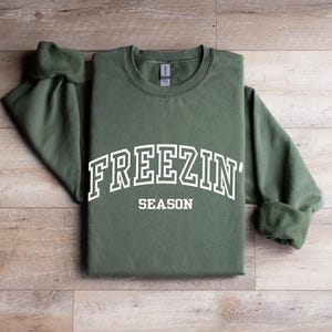Może przedstawiać: Zielona bluza z białym napisem "Freezin' Season".
