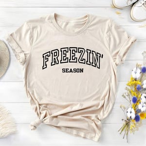 Może przedstawiać: Beżowa koszulka z czarnym napisem "FREEZIN' SEASON".