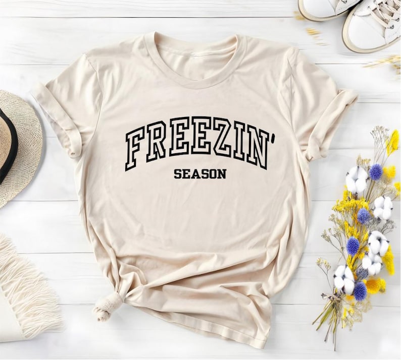 Może przedstawiać: Beżowa koszulka z czarnym napisem "FREEZIN' SEASON".