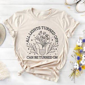May include: A beige t-shirt with a black graphic of a lightbulb with flowers growing out of it. The text "All lights turned off can be turned on" is printed on the shirt.