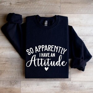 K&ouml;nnte beinhalten: Schwarzer Pullover mit wei&szlig;em Text, der "So Apparently I Have An Attitude" lautet, mit einem kleinen wei&szlig;en Herz unter dem Text.