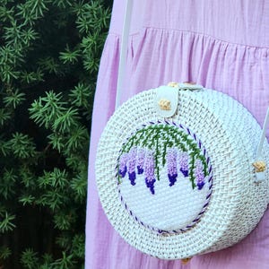 May include: White round woven bag with a white embroidered design of purple flowers and green leaves. The bag has a white strap.