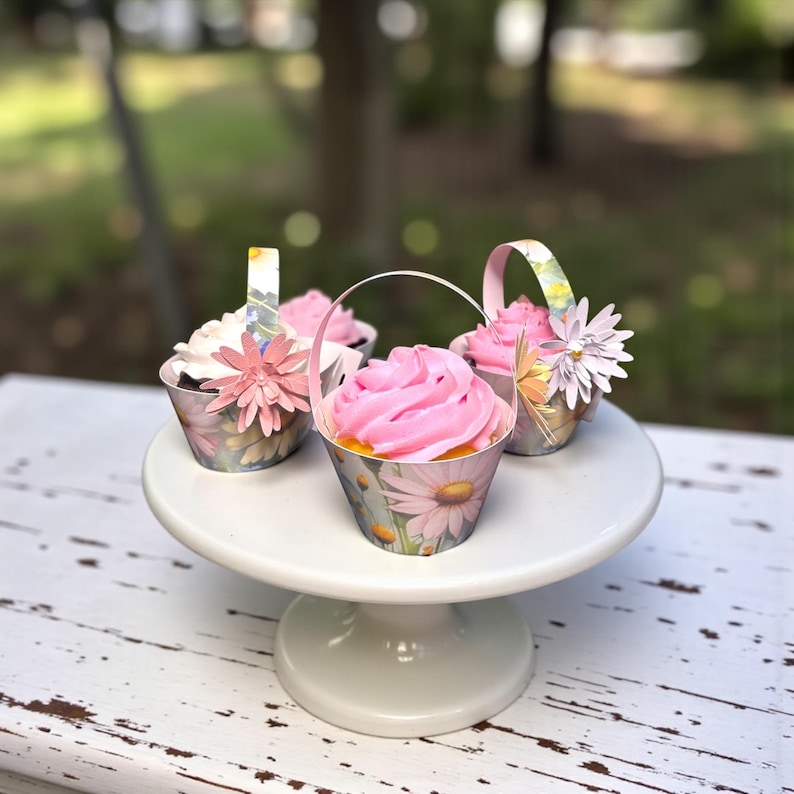 May include: Four cupcakes with pink frosting and floral decorations sit on a white cake stand. The cupcakes are in small metal containers with daisy patterns and paper handles. A few paper flowers are attached to the containers.