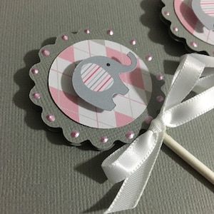 May include: Gray and pink elephant cupcake topper with a white ribbon. The elephant is on a pink and white gingham background.