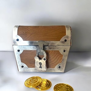 May include: A small, brown wooden treasure chest with silver-colored metal trim and a matching lock. Three gold coins are scattered in front of the chest, suggesting a pirate or adventure theme.