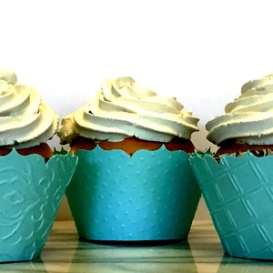 May include: Three cupcakes with white frosting swirls sit in decorative teal paper cups. The cupcake liners have different embossed patterns. The cupcakes are on a reflective surface against a white background.