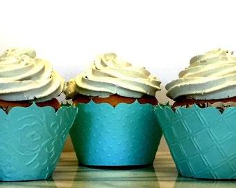 May include: Three cupcakes with white frosting swirls sit in decorative teal paper cups. The cupcake liners have different embossed patterns. The cupcakes are on a reflective surface against a white background.