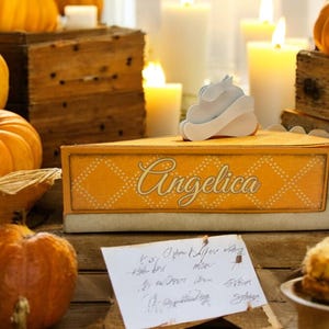 May include: A yellow and white pie box with the word "Angelica" written on it. The box is decorated with a white whipped cream topping and is surrounded by pumpkins and candles.