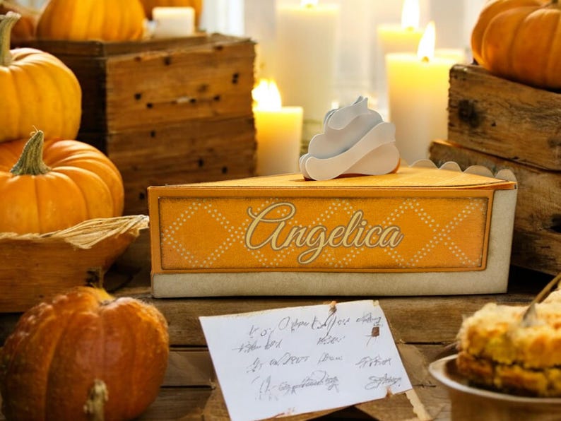 May include: A yellow and white pie box with the word "Angelica" written on it. The box is decorated with a white whipped cream topping and is surrounded by pumpkins and candles.