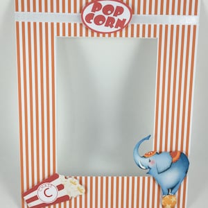 May include: Orange and white striped picture frame with a red and white popcorn sign at the top, a blue elephant with a red and white hat and a red and white popcorn box at the bottom. The frame has a clear opening for a photo.