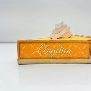 May include: A yellow and white cardboard pie box with a white whipped cream topping. The box has a scalloped edge and says "Angelica" on the front.