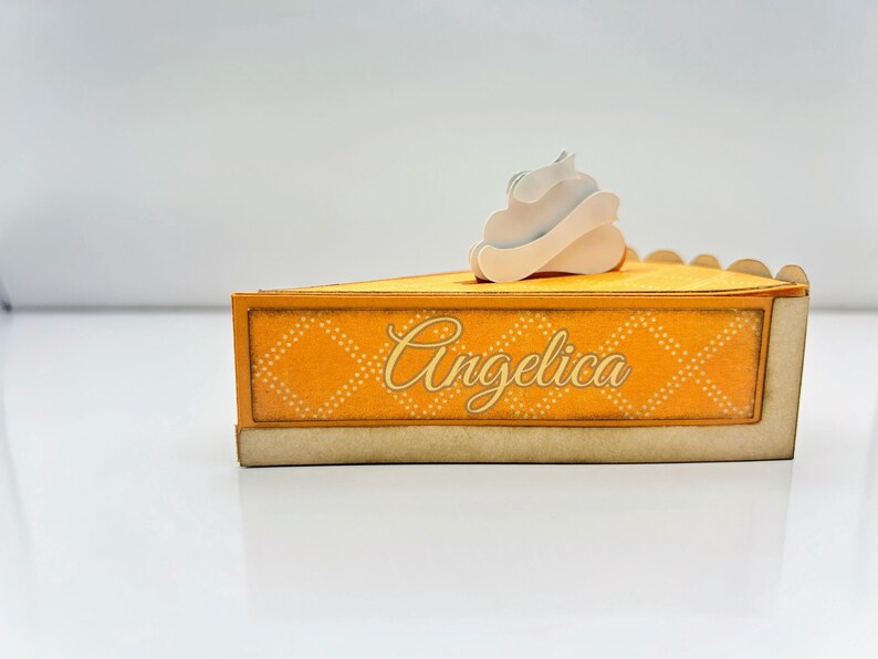 May include: A yellow and white cardboard pie box with a white whipped cream topping. The box has a scalloped edge and says "Angelica" on the front.