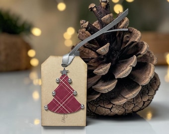 May include: A rectangular tan gift tag featuring a red and white plaid Christmas tree cutout adorned with small silver beads and a star topper. The tag includes a silver ribbon loop and the word Merry printed in script at the bottom.