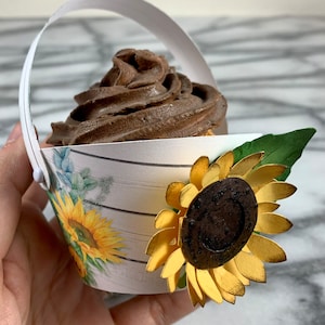 May include: A white paper cupcake holder with a brown chocolate cupcake and a gold sunflower decoration. The holder has a white handle and a woodgrain pattern with a floral design.
