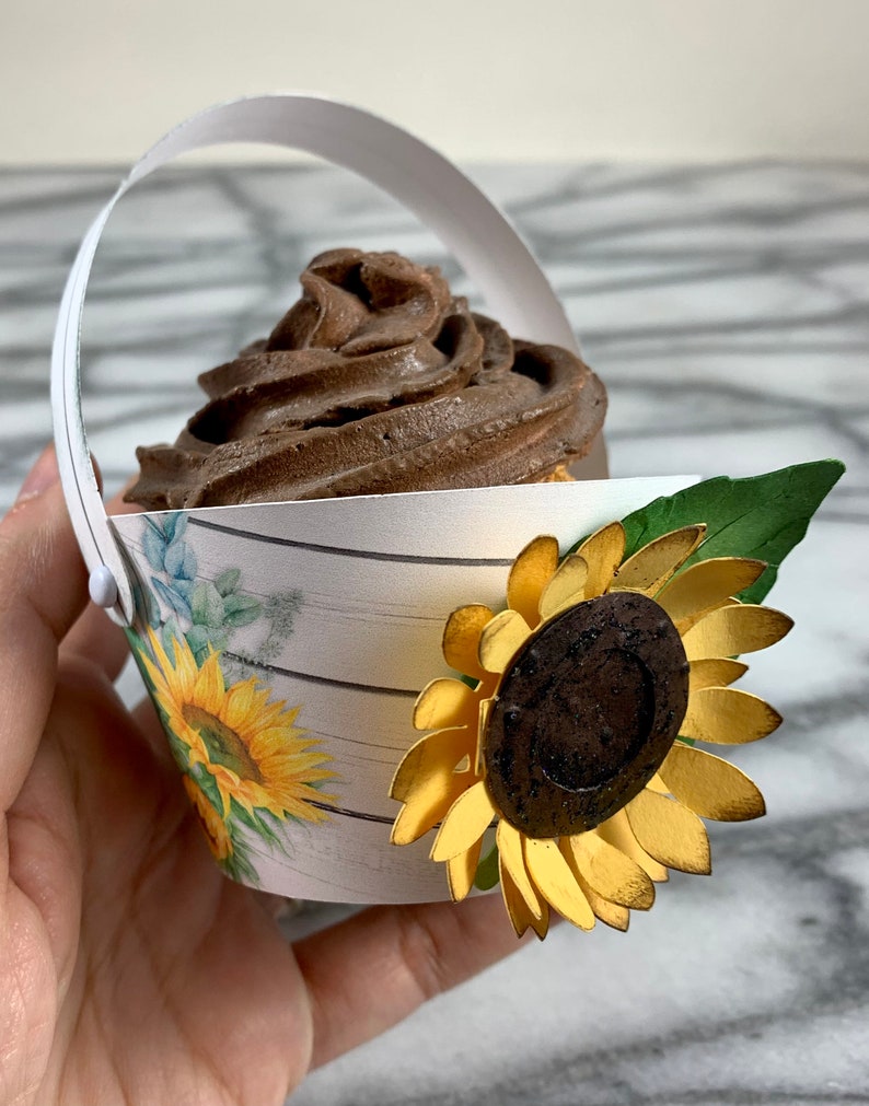 May include: A white paper cupcake holder with a brown chocolate cupcake and a gold sunflower decoration. The holder has a white handle and a woodgrain pattern with a floral design.