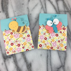 May include: Two small gift bags with a floral pattern. One bag features a bee with brown and yellow stripes and the other features a butterfly with pink and blue wings.