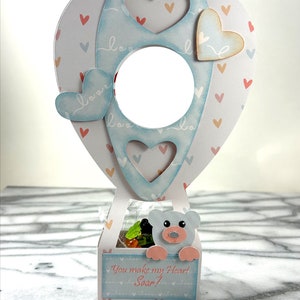 May include: A light blue and white hot air balloon shaped box with a teddy bear peeking out of the top. The box has a heart-shaped opening and is decorated with small hearts and the word "love". The box has a label that says "You make my heart soar!"