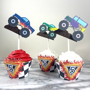 May include: Three monster truck cupcake toppers with a blue, purple, and green monster truck design. The toppers are on cupcakes with black and white checkered cupcake wrappers with a yellow and orange flame design and the number "5" in a blue circle.