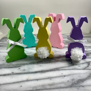May include: Five wooden bunny figurines in different pastel colours. Each bunny has a white pom-pom tail and a white ribbon around its neck.