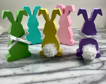May include: Five wooden bunny figurines in different pastel colors. Each bunny has a white pom-pom tail and a white ribbon around its neck.
