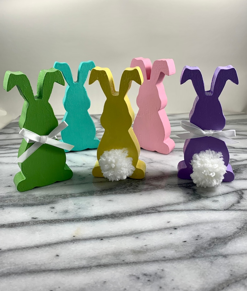 May include: Five wooden bunny figurines in different pastel colours. Each bunny has a white pom-pom tail and a white ribbon around its neck.