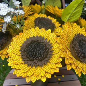 May include: A bouquet of paper sunflowers with yellow petals and dark brown centers. The petals are printed with text. White and yellow accent flowers and green leaves are also included in the arrangement. The bouquet is in a dark green vase.