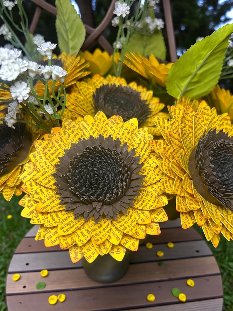 May include: A bouquet of paper sunflowers with yellow petals and dark brown centers. The petals are printed with text. White and yellow accent flowers and green leaves are also included in the arrangement. The bouquet is in a dark green vase.