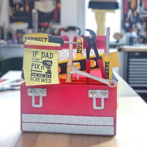 May include: A red toolbox-shaped card with silver accents and a variety of paper tools inside. The tools include a hammer, wrench, and level. A yellow notepad with the text "IF DAD CAN'T FIX IT WE'RE ALL SCREWED" is also visible.