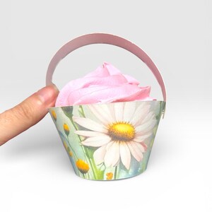 May include: A cupcake in a decorative paper basket with a daisy design. The cupcake has pink frosting. The basket has a handle and is held by a hand.