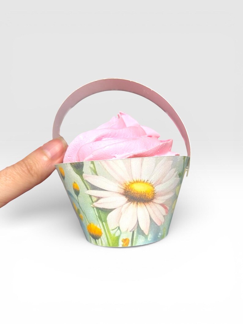 May include: A cupcake in a decorative paper basket with a daisy design. The cupcake has pink frosting. The basket has a handle and is held by a hand.