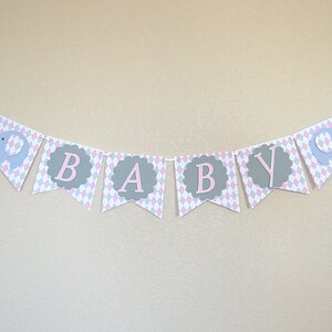 May include: A pink and gray baby banner with the word "BABY" in pink letters on a gray background. The banner has a diamond pattern and features gray elephant cutouts.