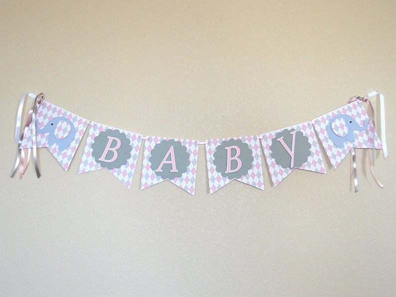 May include: A pink and gray baby banner with the word "BABY" in pink letters on a gray background. The banner has a diamond pattern and features gray elephant cutouts.