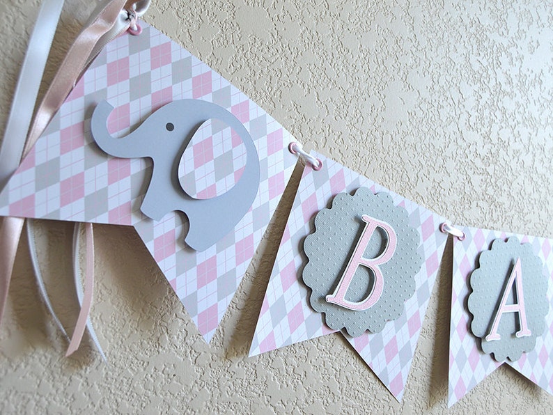 May include: A pink and gray baby banner with the word "BABY" in pink letters on a gray background. The banner is decorated with a gray elephant and a pink and gray checkered pattern.