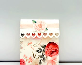 May include: A white favor bag with a floral design featuring pink and red roses, green leaves, and a pink flower with a white heart-shaped border.