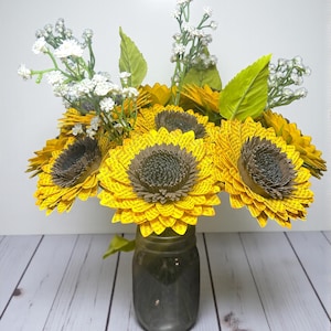 May include: A bouquet of paper sunflowers in a gray mason jar. The sunflowers have yellow petals and brown centers. White baby's breath and green leaves are interspersed among the sunflowers. The jar sits on a white wooden surface.