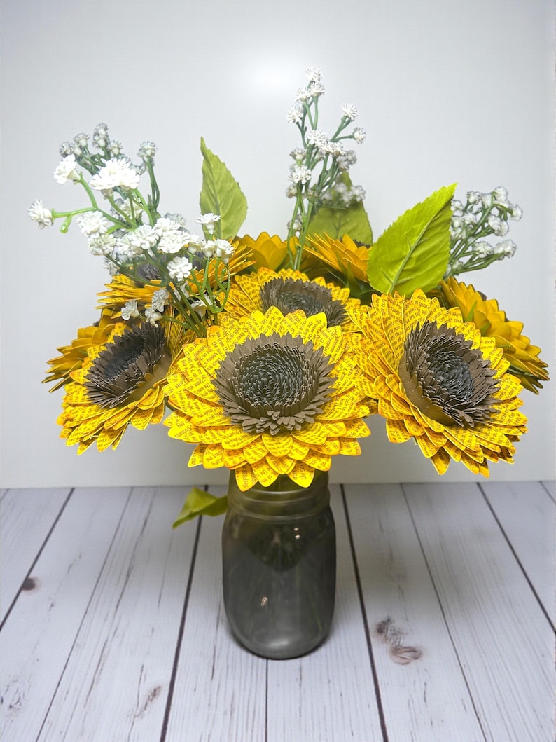 May include: A bouquet of paper sunflowers in a gray mason jar. The sunflowers have yellow petals and brown centers. White baby's breath and green leaves are interspersed among the sunflowers. The jar sits on a white wooden surface.