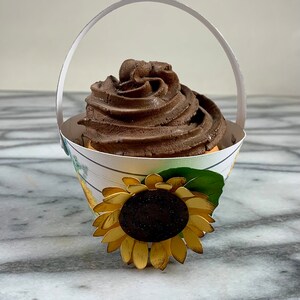 May include: A chocolate cupcake with a swirl of frosting in a white paper cupcake holder with a sunflower design and a handle.