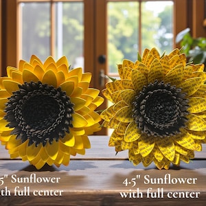 May include: Two paper sunflowers with full centers are displayed on a wooden surface. The sunflower on the left is 3.5 inches in diameter, and the one on the right is 4.5 inches. Both have yellow petals and dark centers.
