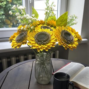 May include: A bouquet of paper sunflowers in a clear glass jar, with white filler flowers and green leaves. The sunflowers are yellow with brown centers. A black mug and an open book sit on a dark wooden table near a window.