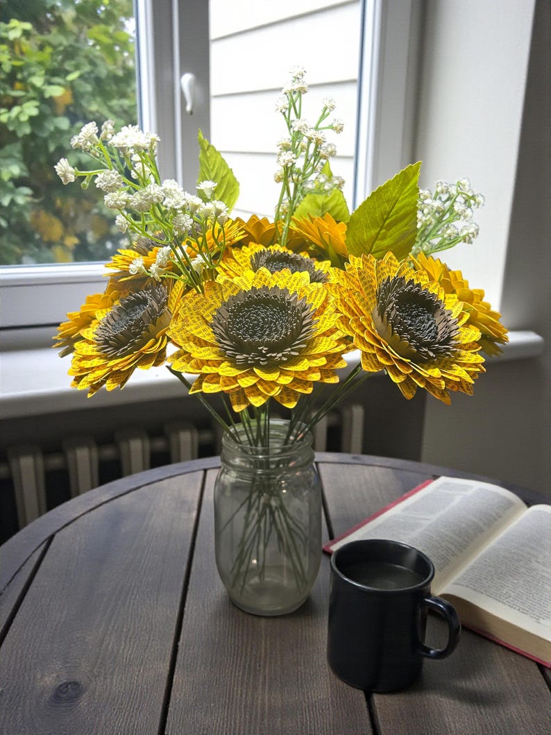 May include: A bouquet of paper sunflowers in a clear glass jar, with white filler flowers and green leaves. The sunflowers are yellow with brown centers. A black mug and an open book sit on a dark wooden table near a window.
