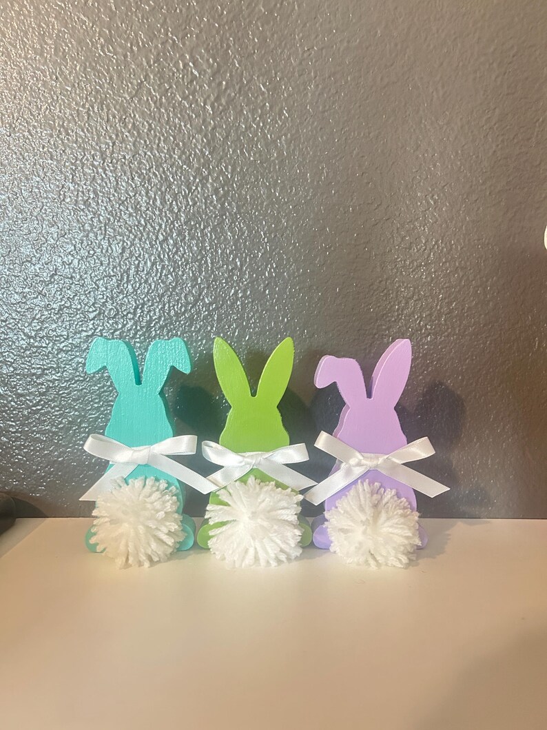 May include: Three wooden bunny figurines, painted in pastel colours of teal, green, and purple. Each bunny has a white bow and a white pom-pom tail.