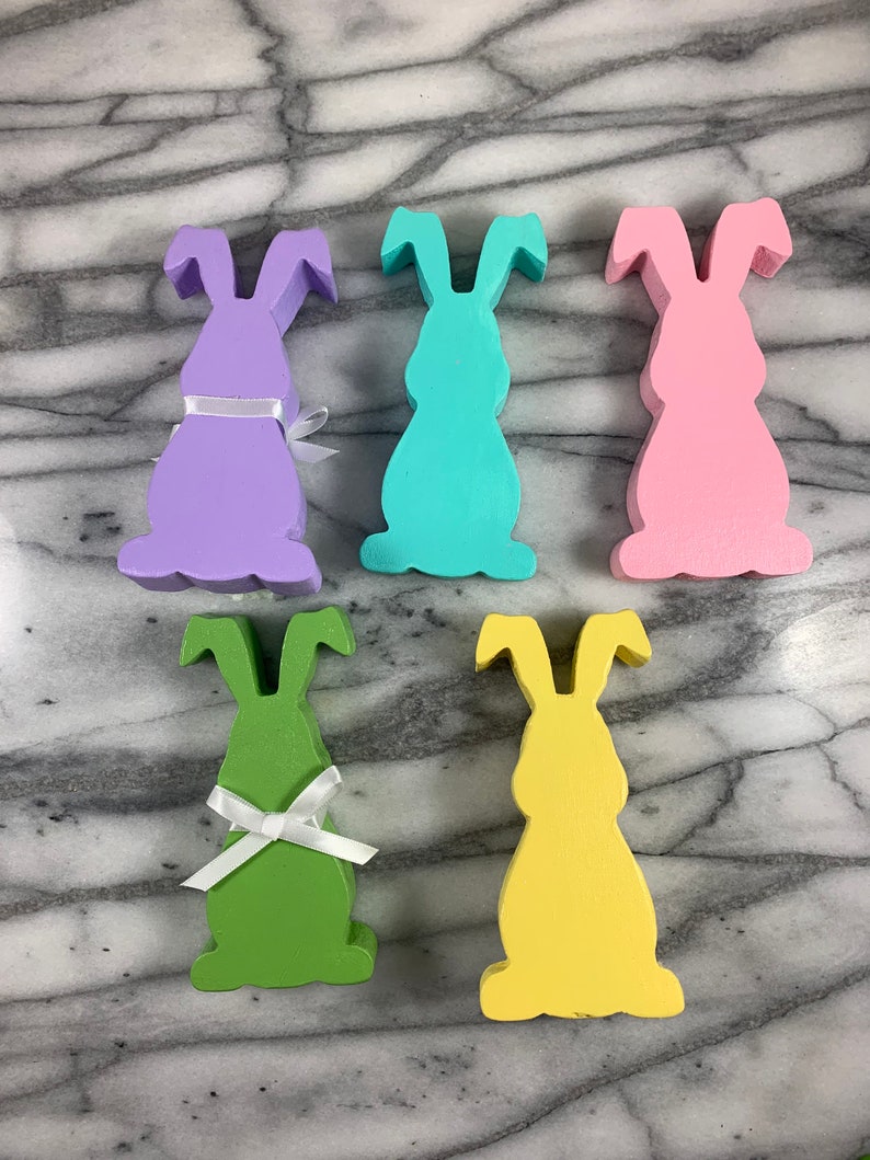 May include: Five wooden bunny rabbit figurines in different pastel colours. The bunnies are all the same size and shape, and they have white ribbon bows around their necks. The bunnies are arranged in a row on a marble surface.