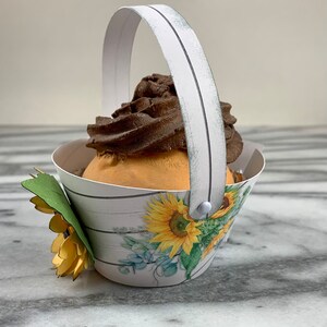 May include: A white paper cupcake holder with a white handle and a sunflower design. The holder is filled with a chocolate cupcake with a swirl of frosting.