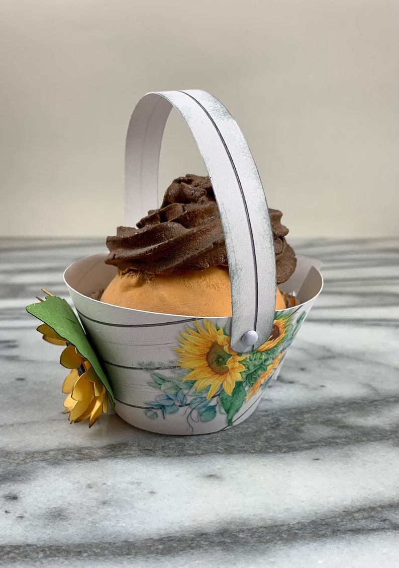 May include: A white paper cupcake holder with a white handle and a sunflower design. The holder is filled with a chocolate cupcake with a swirl of frosting.