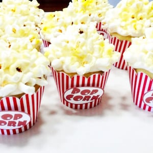 May include: Close-up of cupcakes decorated with white frosting and popcorn, resembling popcorn balls. The cupcakes are in red and white striped wrappers with the words "Pop Corn" printed on them.