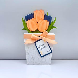 May include: A paper flower arrangement in a white pot with a peach ribbon and a tag that says "Happy Birthday!". The arrangement features orange and navy blue paper tulips with green leaves. The pot has a floral pattern.