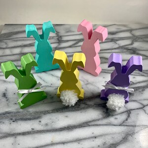 May include: Five wooden bunny figurines painted in pastel colours. The bunnies are standing on a marble surface. Each bunny has a white pom-pom tail and a white ribbon around its neck.