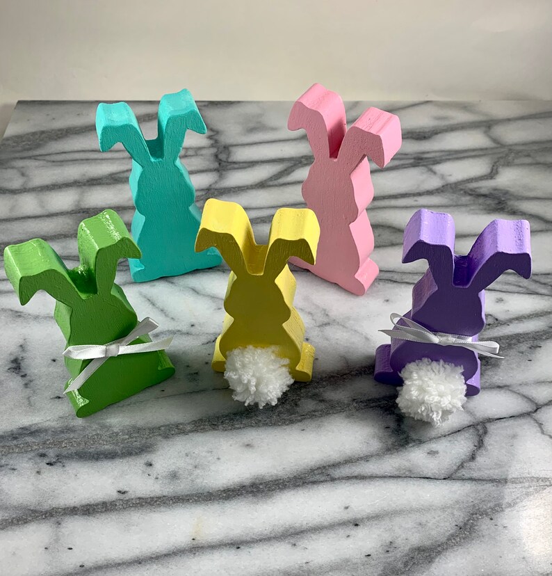 May include: Five wooden bunny figurines painted in pastel colours. The bunnies are standing on a marble surface. Each bunny has a white pom-pom tail and a white ribbon around its neck.